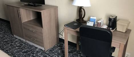 A hotel room with a wooden desk, a black office chair, a television, a lamp, a coffee maker, and a small table with a sign.