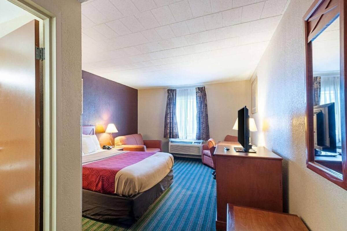 Una habitación de hotel con cama, escritorio, silla, televisor y ventana con cortinas.