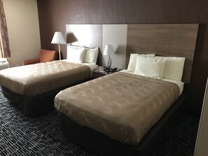 A hotel room with two beds, a nightstand, a lamp, and a chair.