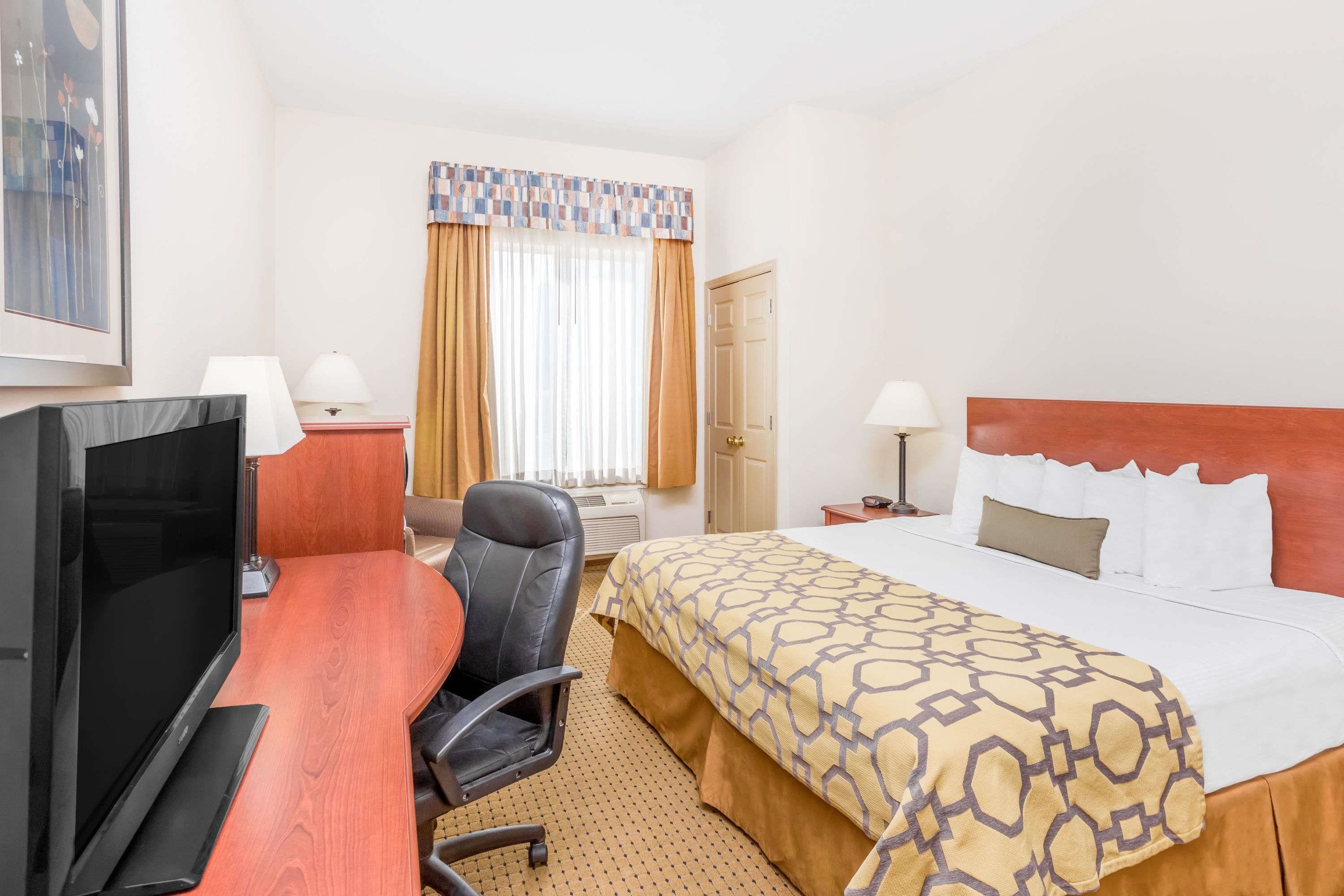 A hotel room with a bed, a desk with a TV, a chair, a window with curtains, and a door.