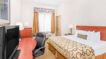 A hotel room with a bed, a desk with a TV, a chair, a window with curtains, and a door.