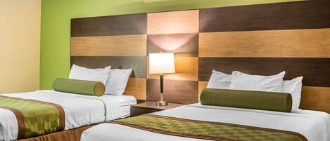 A hotel room with two beds, each with white linens and green throw pillows. There is a nightstand with a lamp between the beds.