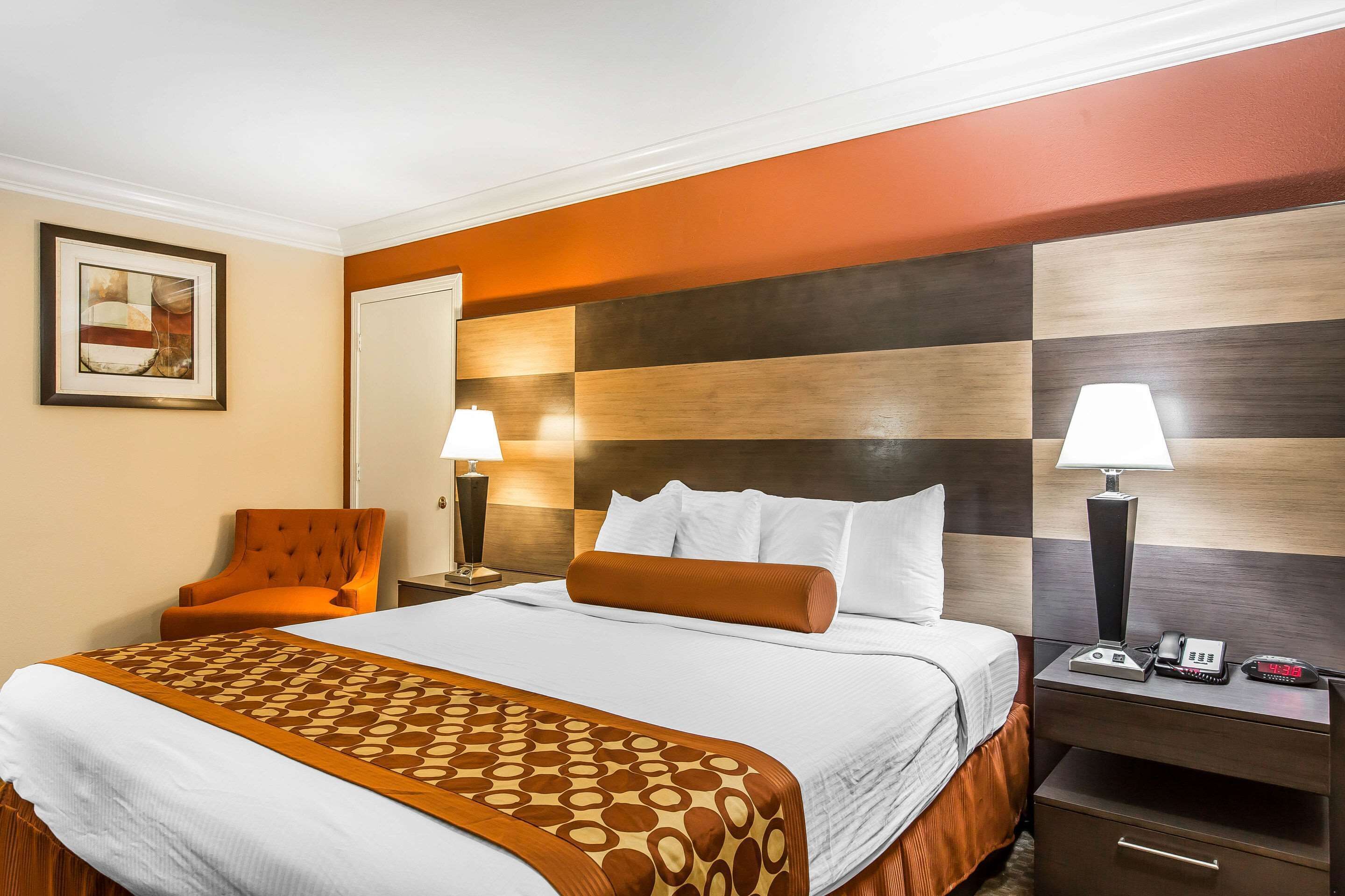 Quarto de hotel com uma cama grande, mesa de cabeceira com abajur, uma cadeira e um quadro na parede.