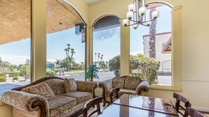 A dining area with a sofa and chairs, a glass table, and large windows with a view of palm trees and a building. - Rodeway Inn & Suites Canyon Lake-Menifee West (Menifee)