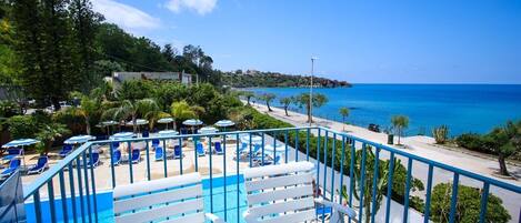 Un área de piscina con sillas reclinables blancas y sombrillas, un cielo azul despejado y vista al mar.