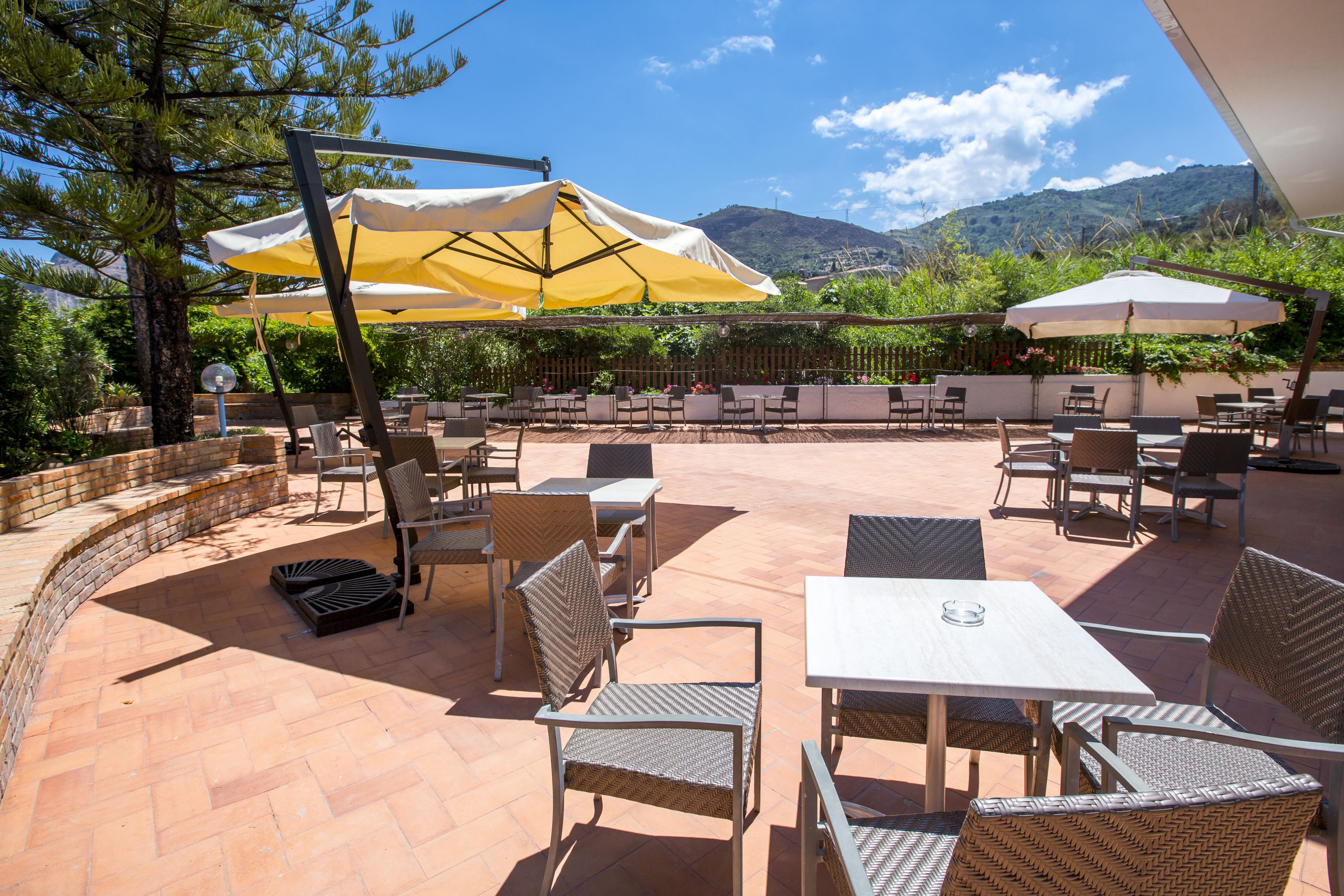 An outdoor patio with wicker furniture and umbrellas, surrounded by a brick wall and greenery, overlooking a mountainous landscape.