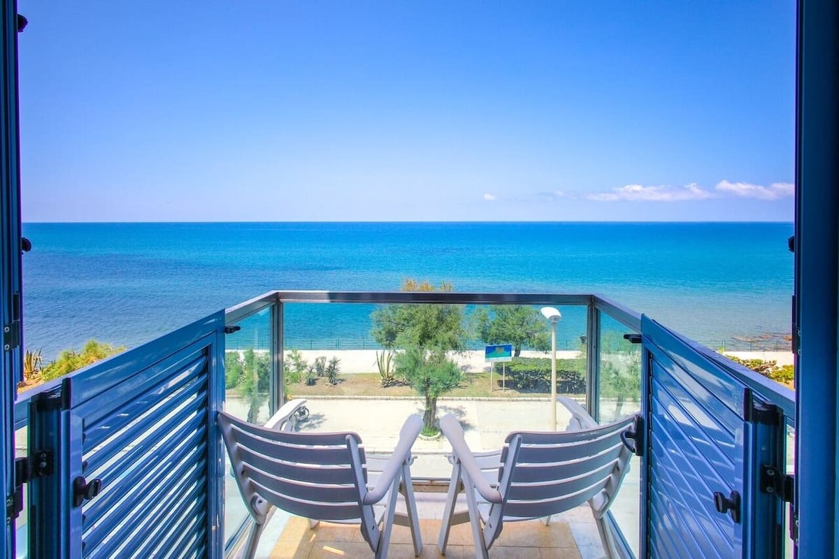 A balcony with two chairs facing a clear blue sea and a sandy beach.