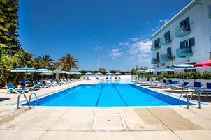Piscina com espreguiçadeiras e guarda-sóis, cercada por palmeiras e um edifício com varandas.