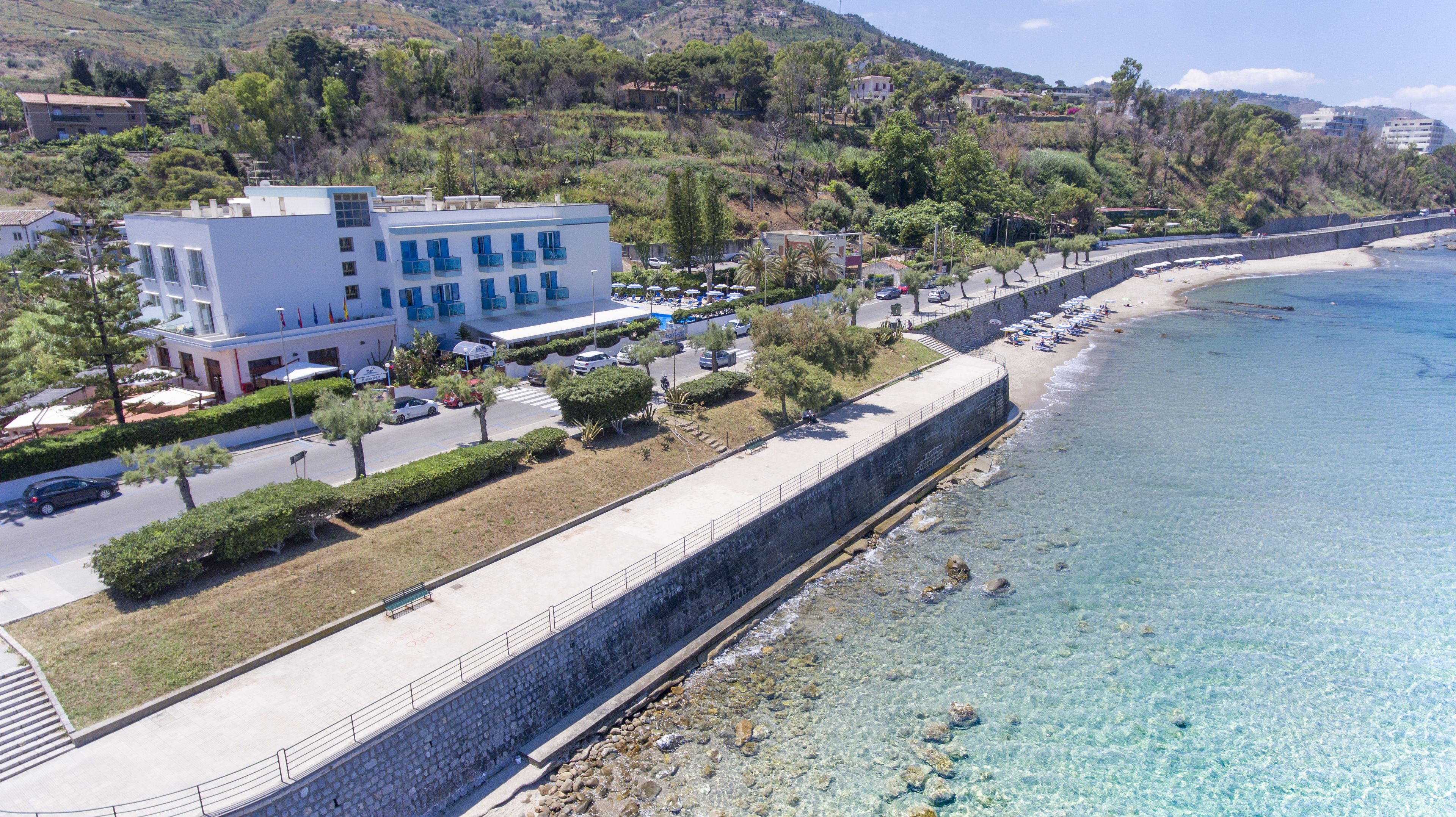 Un hôtel situé sur la côte, doté d’une promenade pavée longeant la plage, d’une eau d’un bleu cristallin et d’un mur de pierre qui sépare la promenade de la plage.