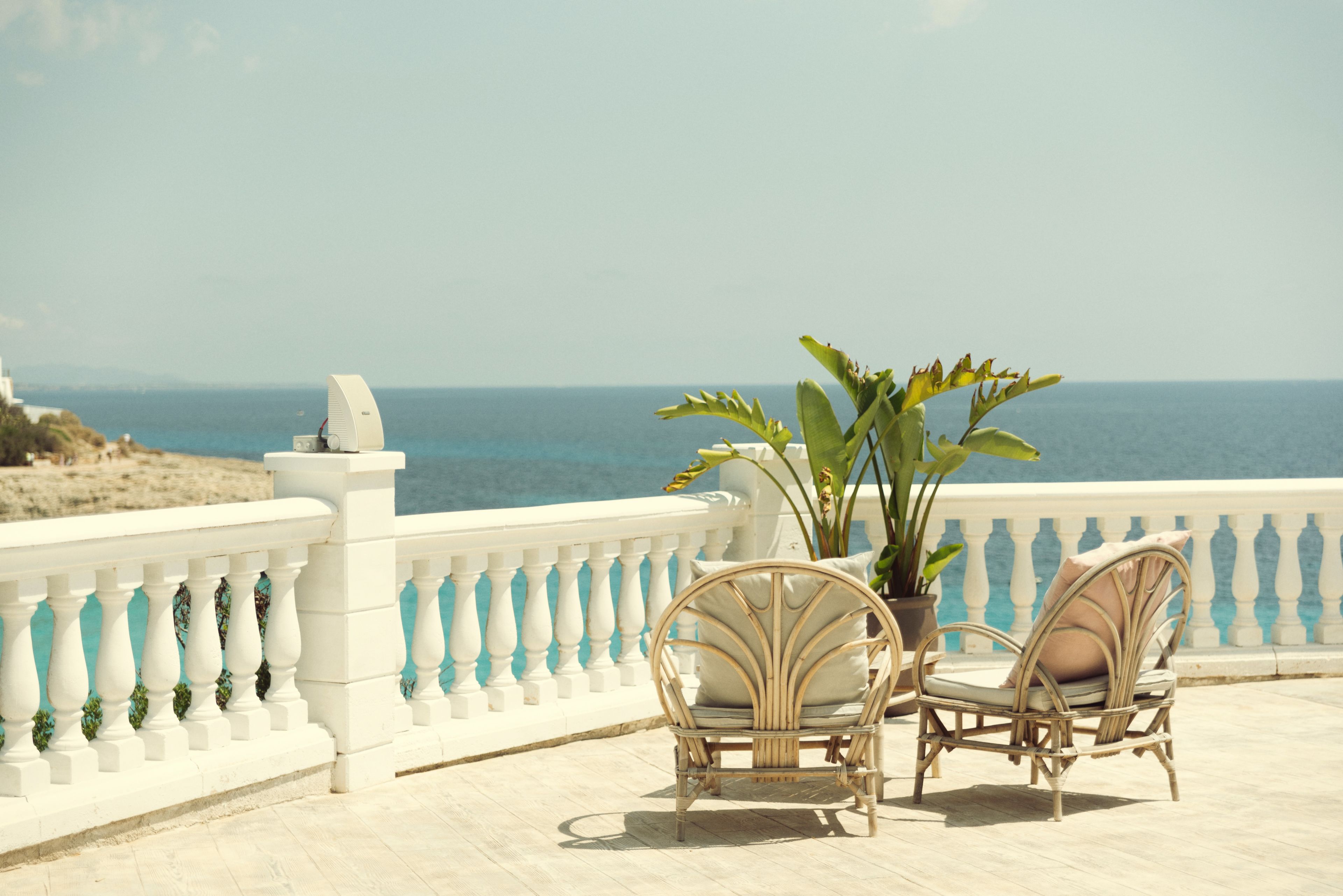 Un balcon avec deux fauteuils en osier et un pot de fleurs, d’où l’on a vue sur une plage et la mer.
