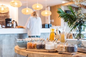Een ontbijtbuffet met diverse etenswaren en drankjes op een houten tafel, met hangende lampen erboven.