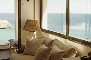 A room with a large window offering a view of the sea and a rocky outcrop. There is a sofa with cushions and a lamp on a side table.