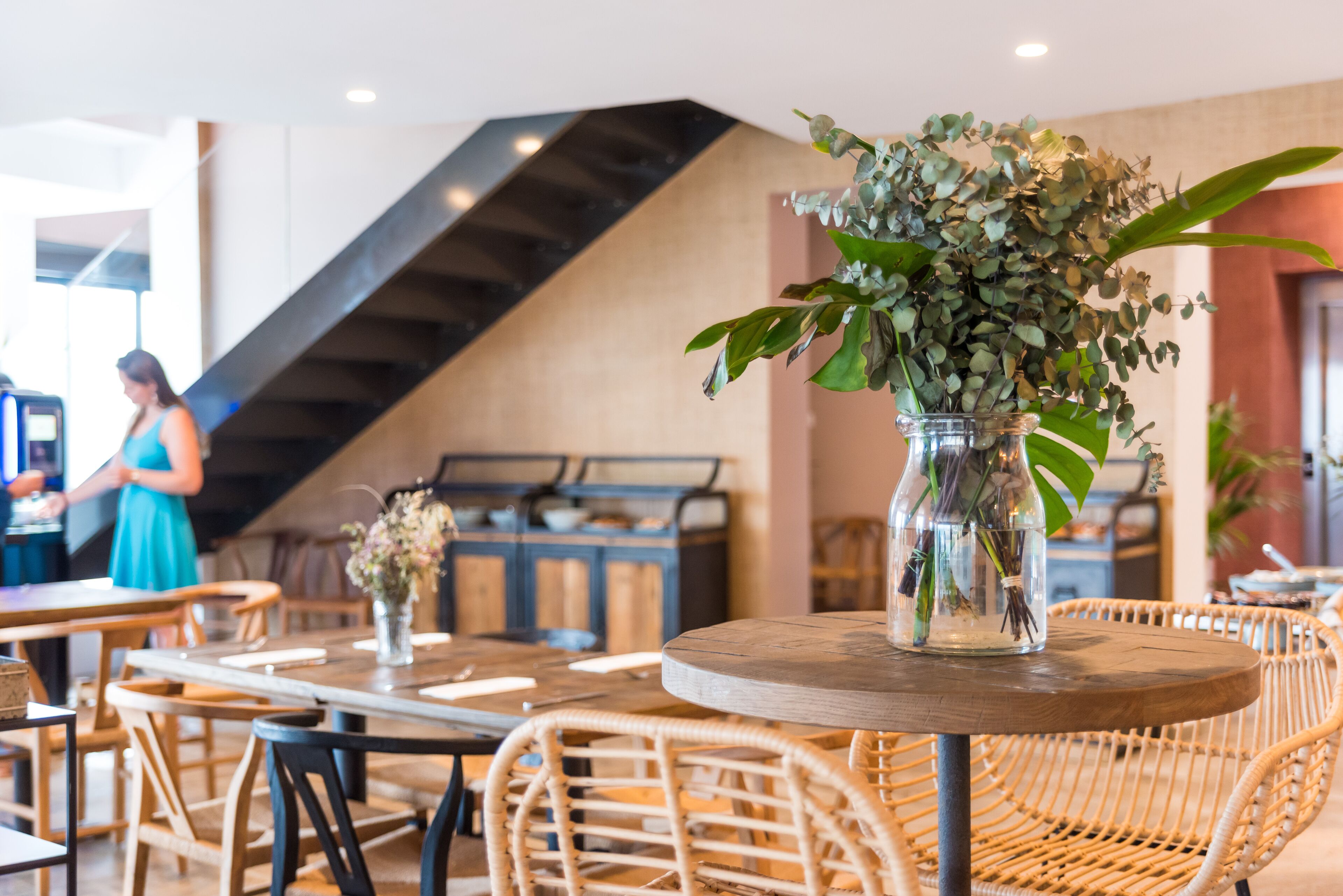 A restaurant with wicker chairs, a round wooden table with a vase of flowers, and a staircase in the background.
