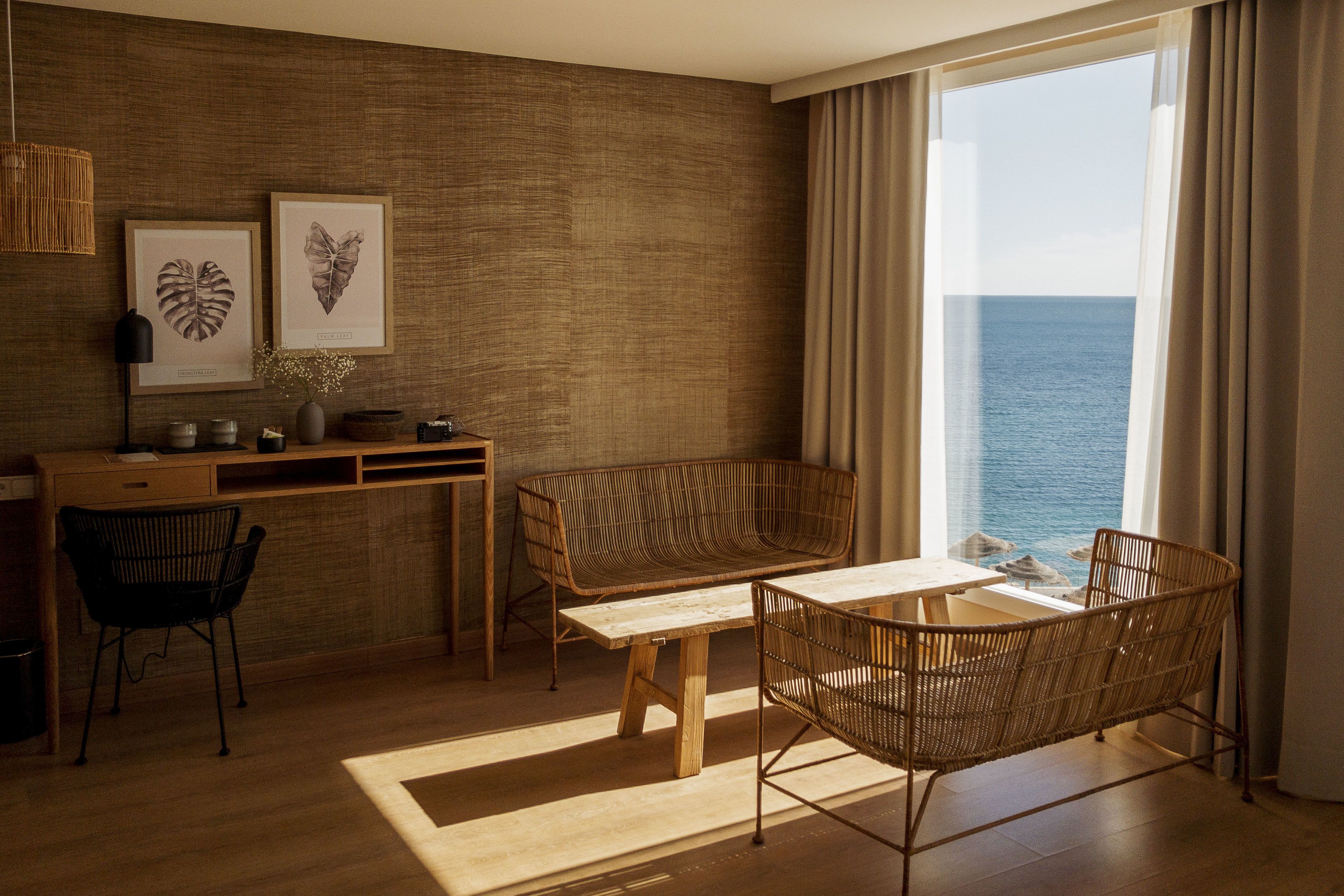 A modern room with a wooden desk, two wicker chairs, a sofa, and a view of the sea.