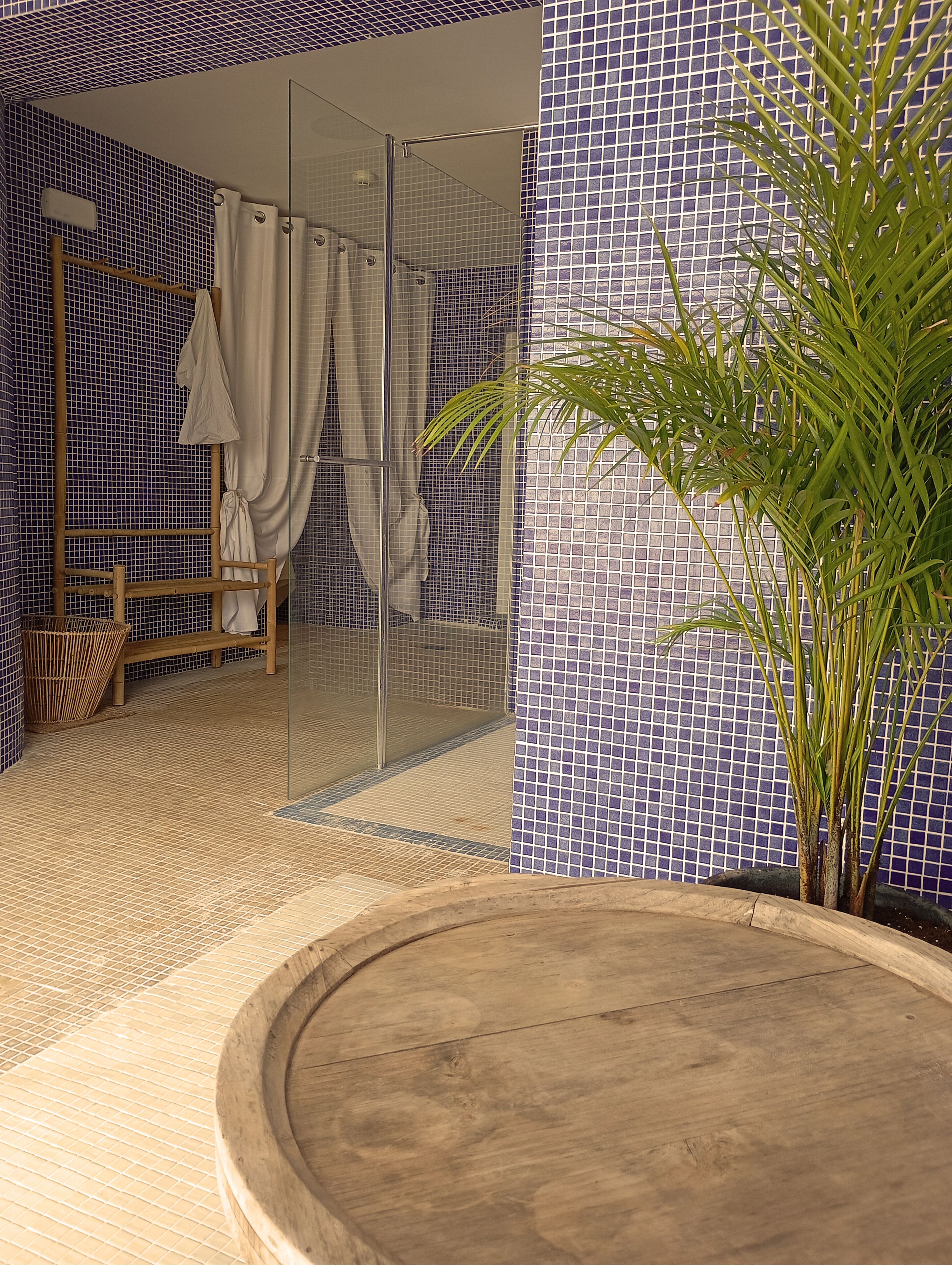 A bathroom with a glass shower enclosure, a wooden bench, a potted plant, and a tiled floor.