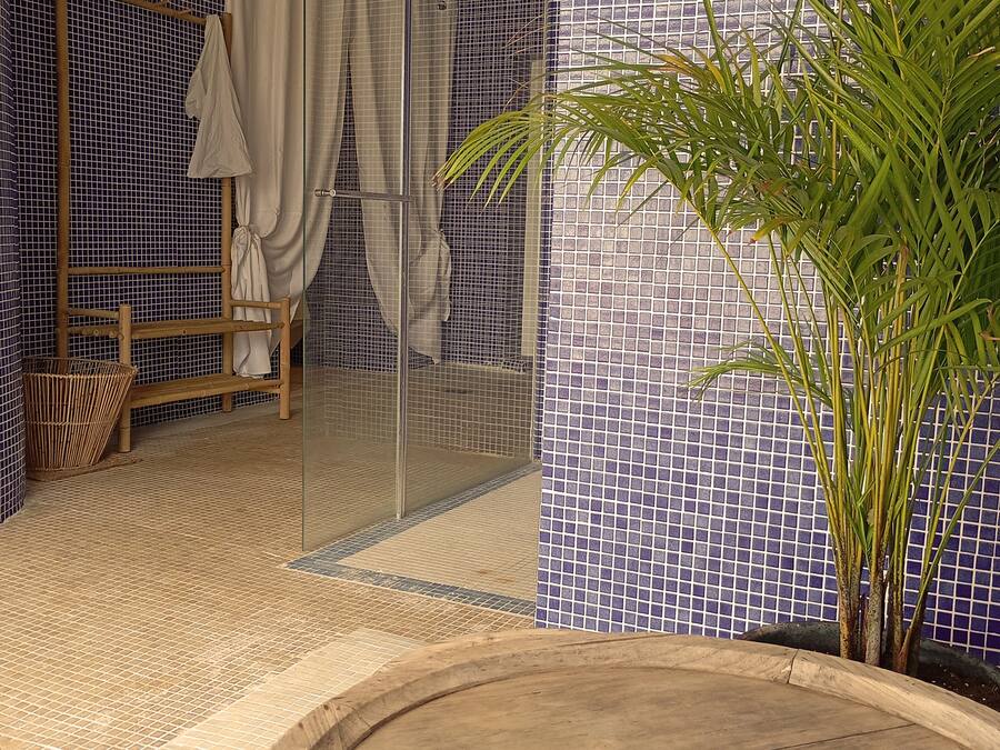 A bathroom with a glass shower enclosure, a wooden bench, a potted plant, and a tiled floor.