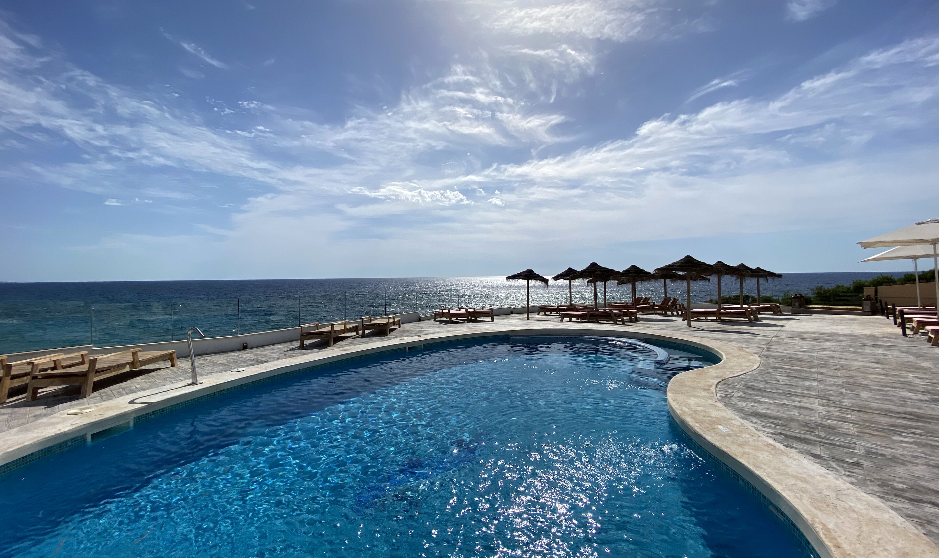 A pool with a curved edge, wooden lounge chairs, and umbrellas, overlooking a clear blue sea under a partly cloudy sky.