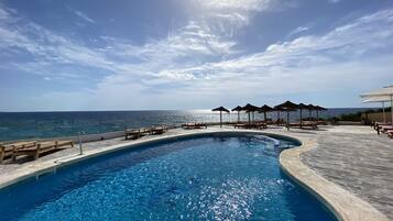 Ein Pool mit geschwungener Kante, Holzliegen und Sonnenschirmen mit Blick auf das klare, blaue Meer unter einem teils bewölkten Himmel.