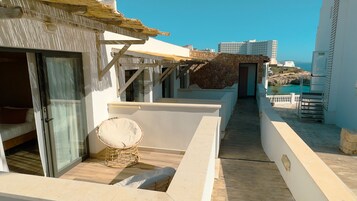 Un balcón estrecho con una silla de mimbre, una mesita y vista al mar y edificios al fondo.
