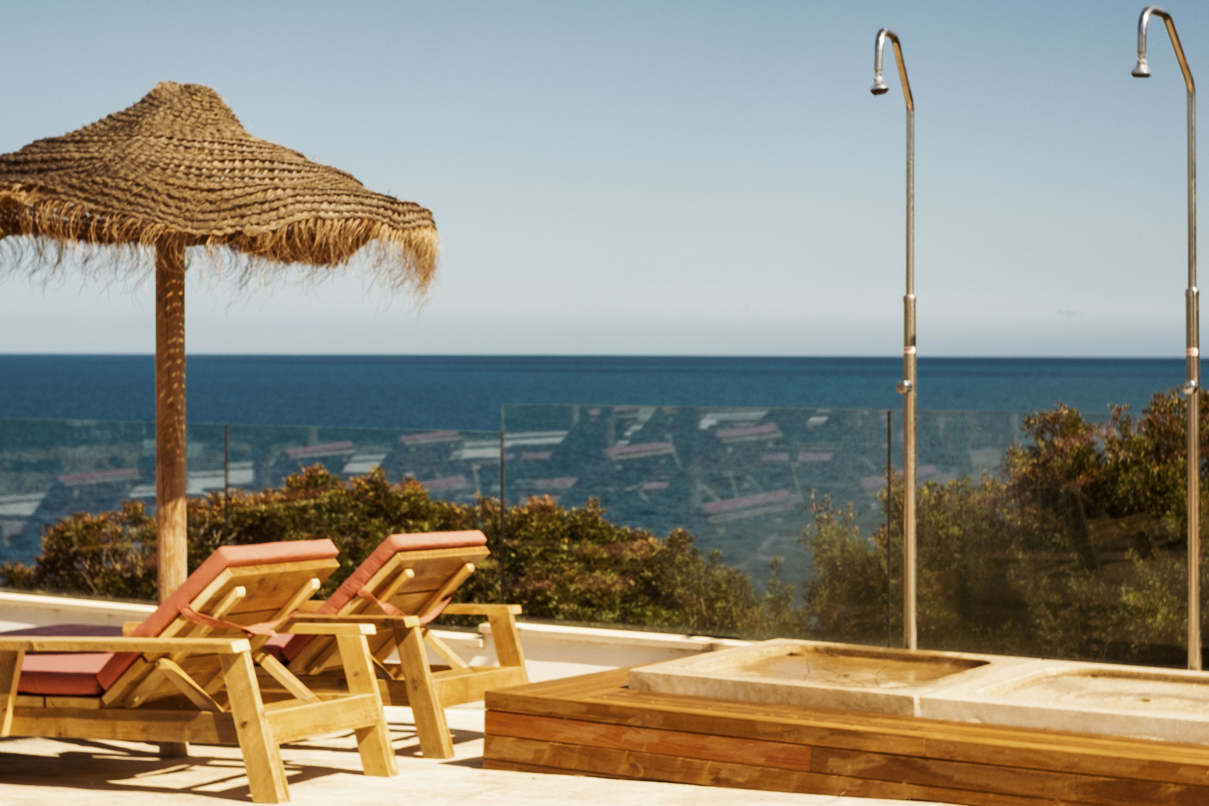 Zona de piscina con sillones de madera y una sombrilla de paja, con vista a un paisaje marino.