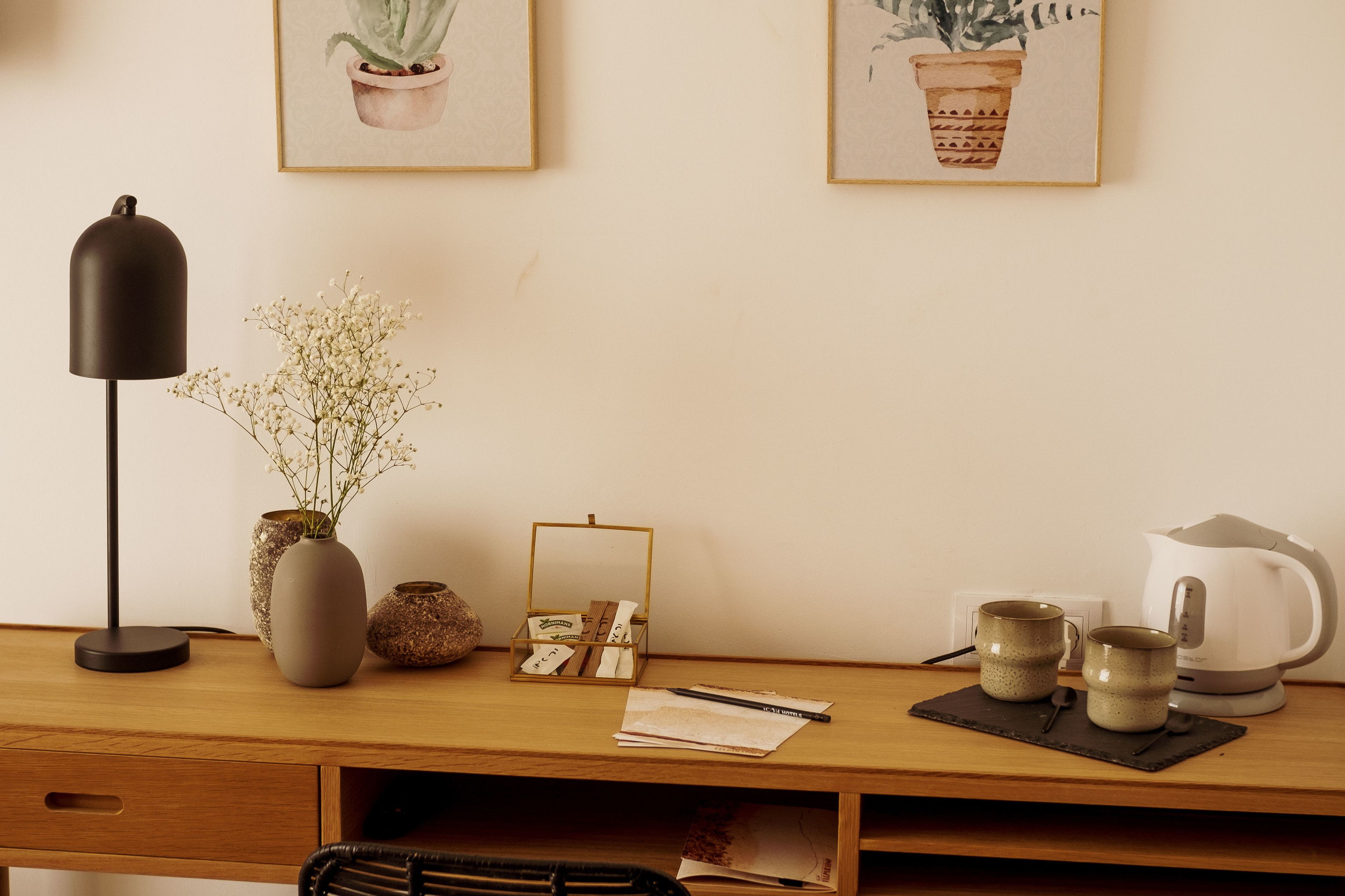 Een houten bureau met een lamp, een vaas met bloemen, een zak tissues, twee mokken en een waterkoker.