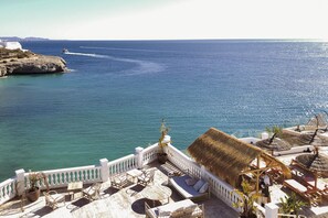 Une terrasse en bord de mer, dotée de sièges, recouverte d’une toiture de chaume et offrant une vue sur la mer.