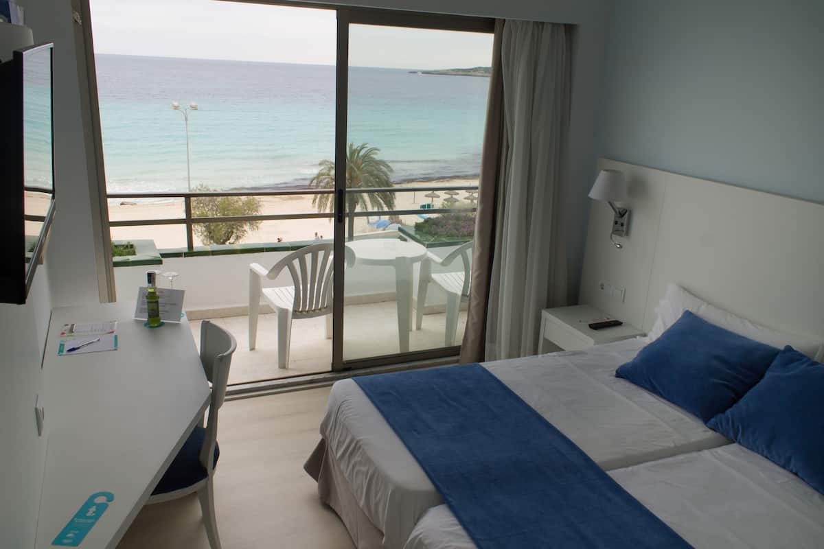 Ein Hotelzimmer mit einem großen Bett, einem Schreibtisch mit Stuhl, einem Fernseher und einem Balkon mit Blick auf Strand und Meer.
