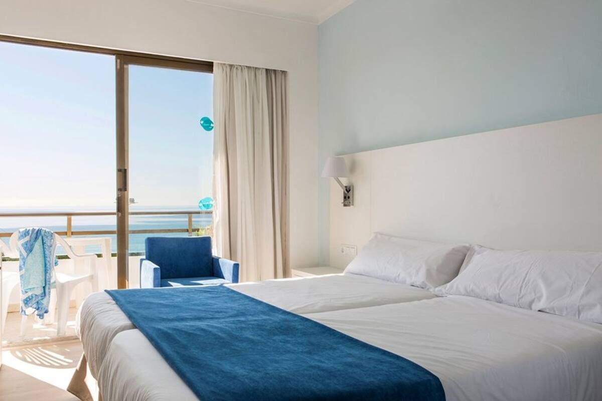 Ein Hotelzimmer mit einem großen Bett, einem Sessel, einem Tisch und Meerblick durch eine Schiebetür.