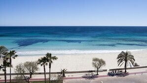 A beach view with clear blue water, sandy shore, and palm trees. - Hotel Anba Romani (Sant Llorenc des Cardassar)