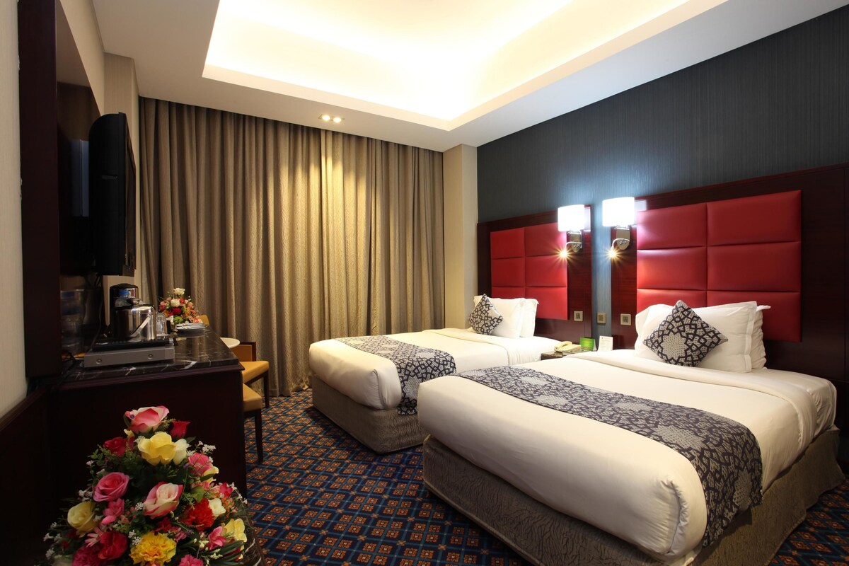 Habitación de hotel con dos camas, un escritorio, televisor de pantalla plana, cafetera y un arreglo floral.