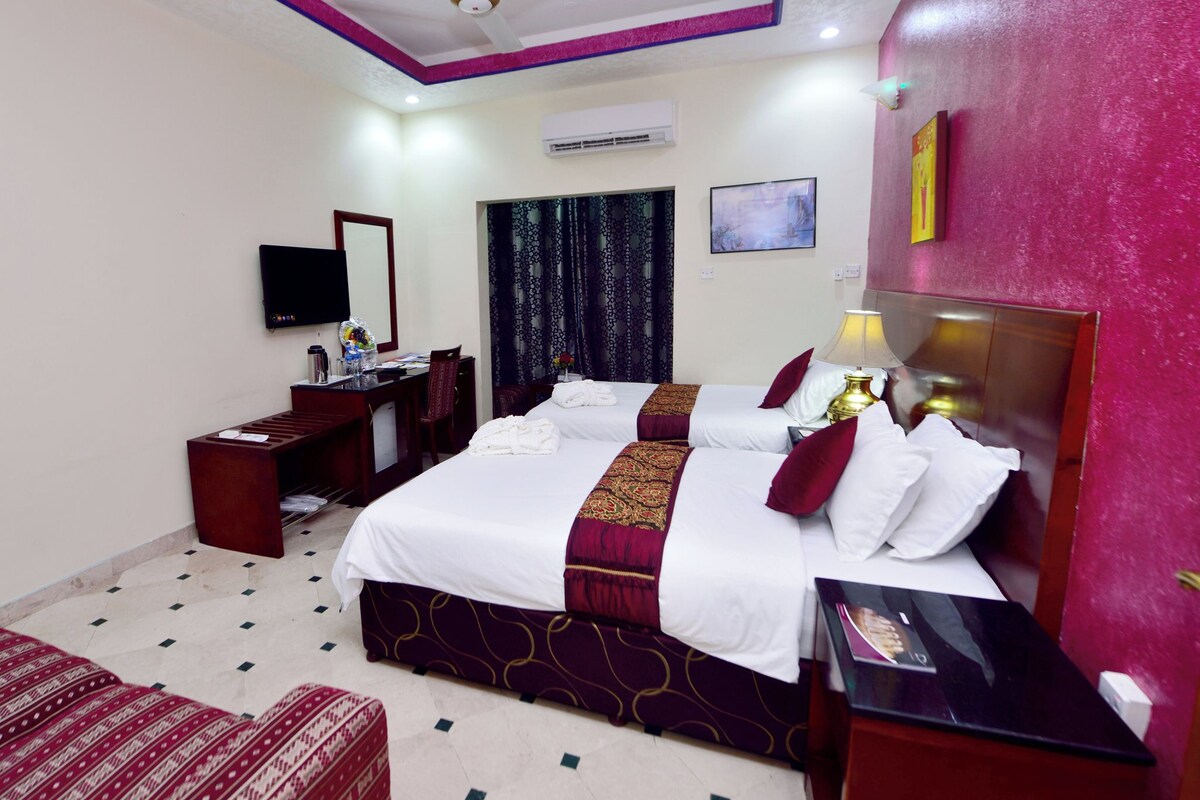 A hotel room with two beds, a desk, a chair, a TV, and a ceiling fan.