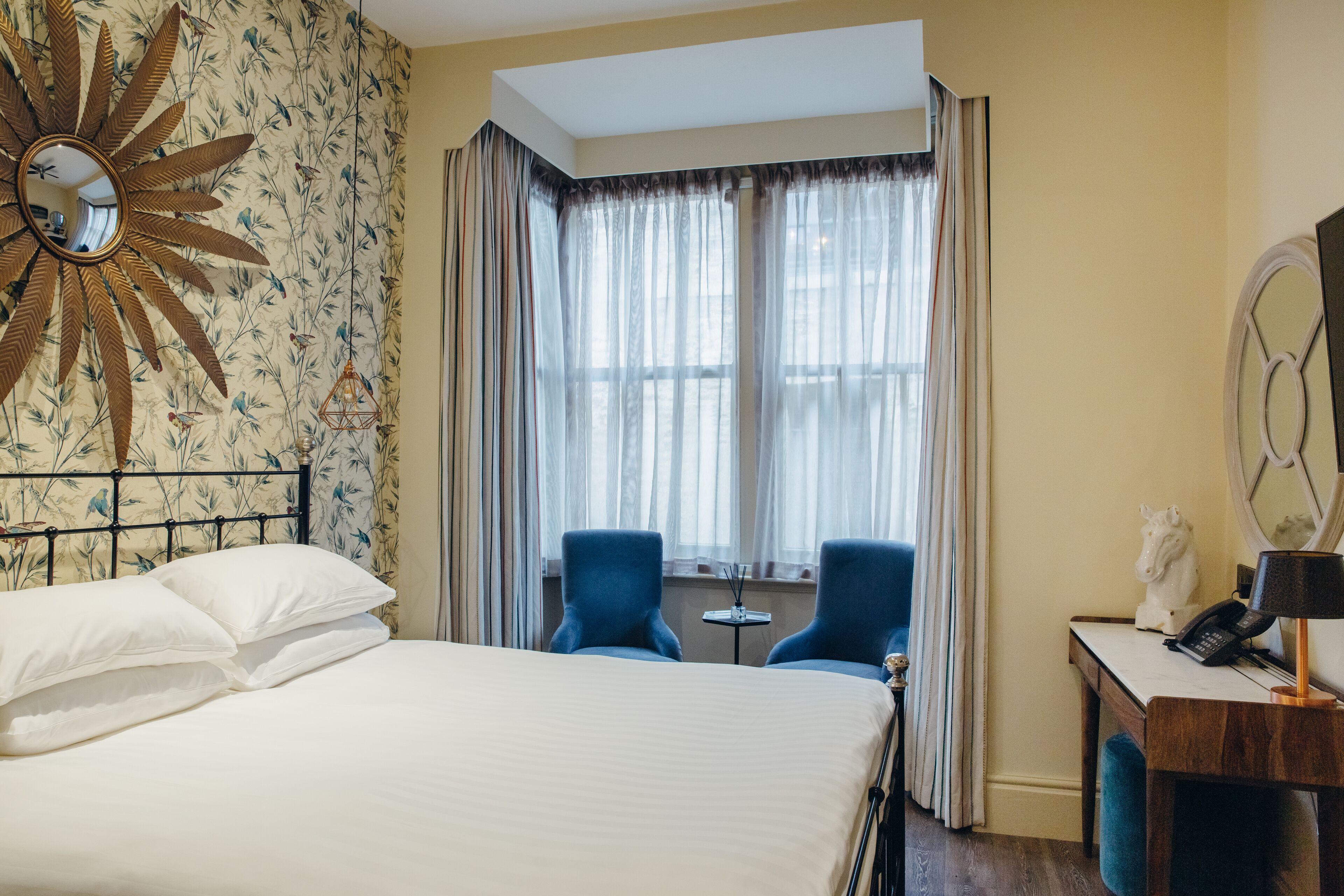 A hotel room with a bed, two blue chairs, a small table, a mirror, and a decorative sunburst wall piece.