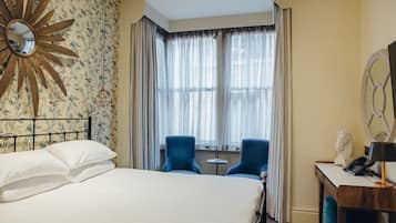 A hotel room with a bed, two blue chairs, a small table, a mirror, and a decorative sunburst wall piece.