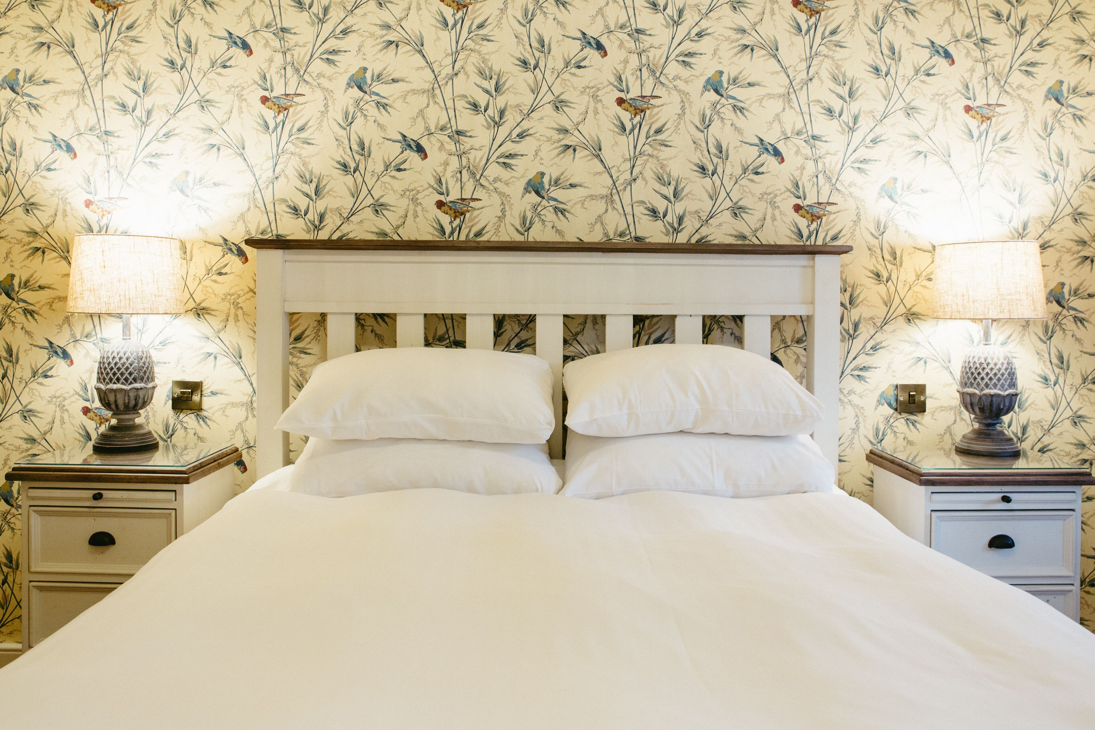 A neatly made bed with white linens, flanked by two nightstands with lamps, set against a floral wallpaper backdrop.