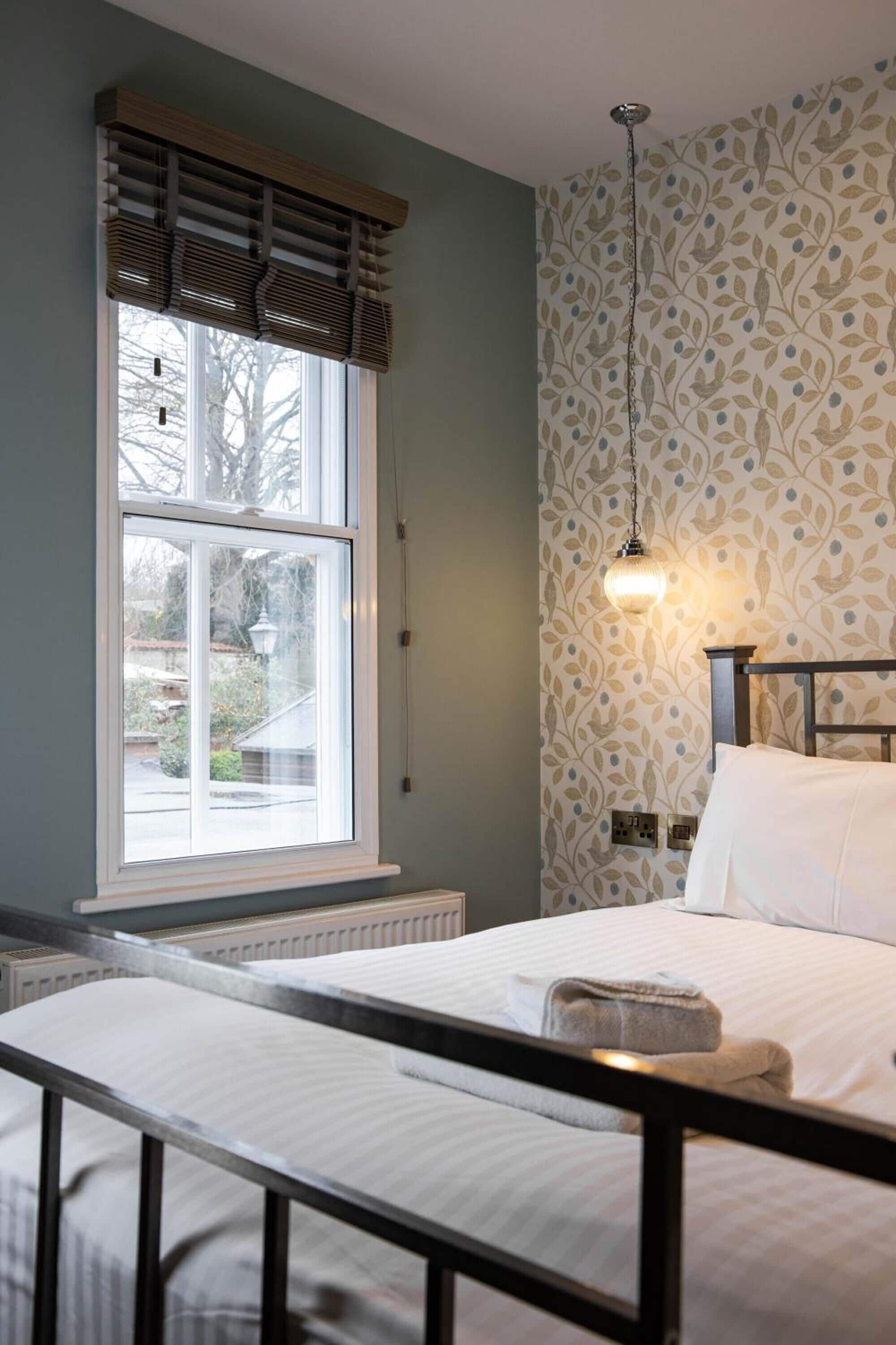 A bedroom with a large window, a bed with white linens, a hanging light fixture, and a patterned wallpaper.