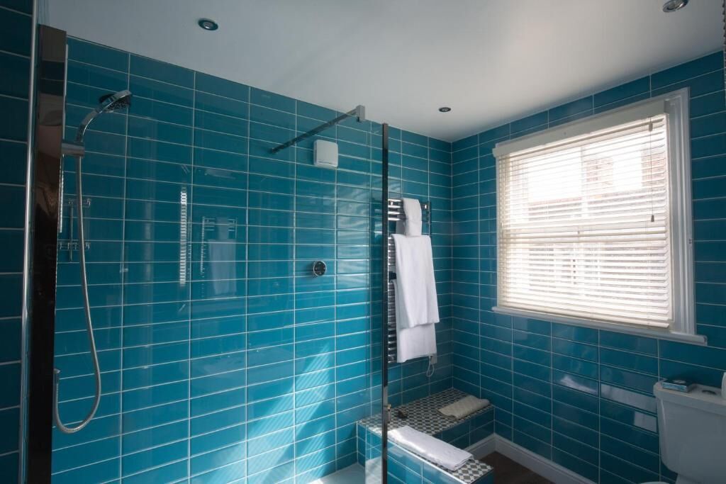 A bathroom with blue tiles, a glass shower enclosure, a toilet, and a window with blinds.