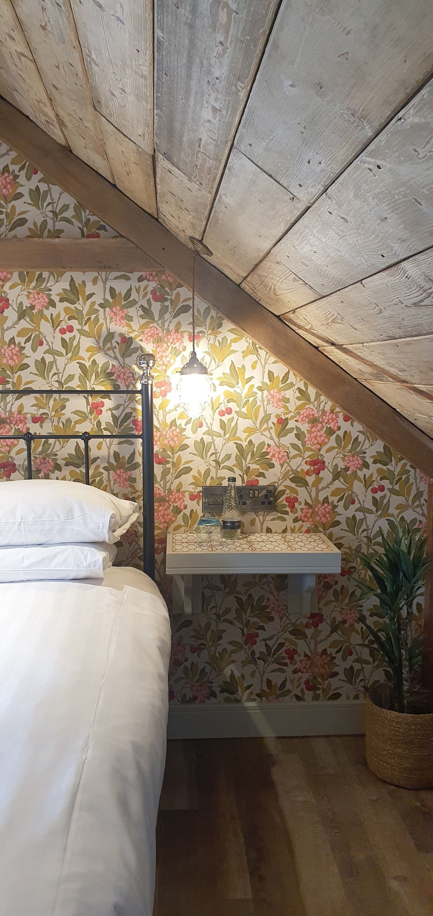 Ein Schlafzimmer mit schrägen Wänden, Blumenpapier, einem Bett mit weißen Leinen, einem Nachttisch mit Lampe und einem Topfpflanzen.