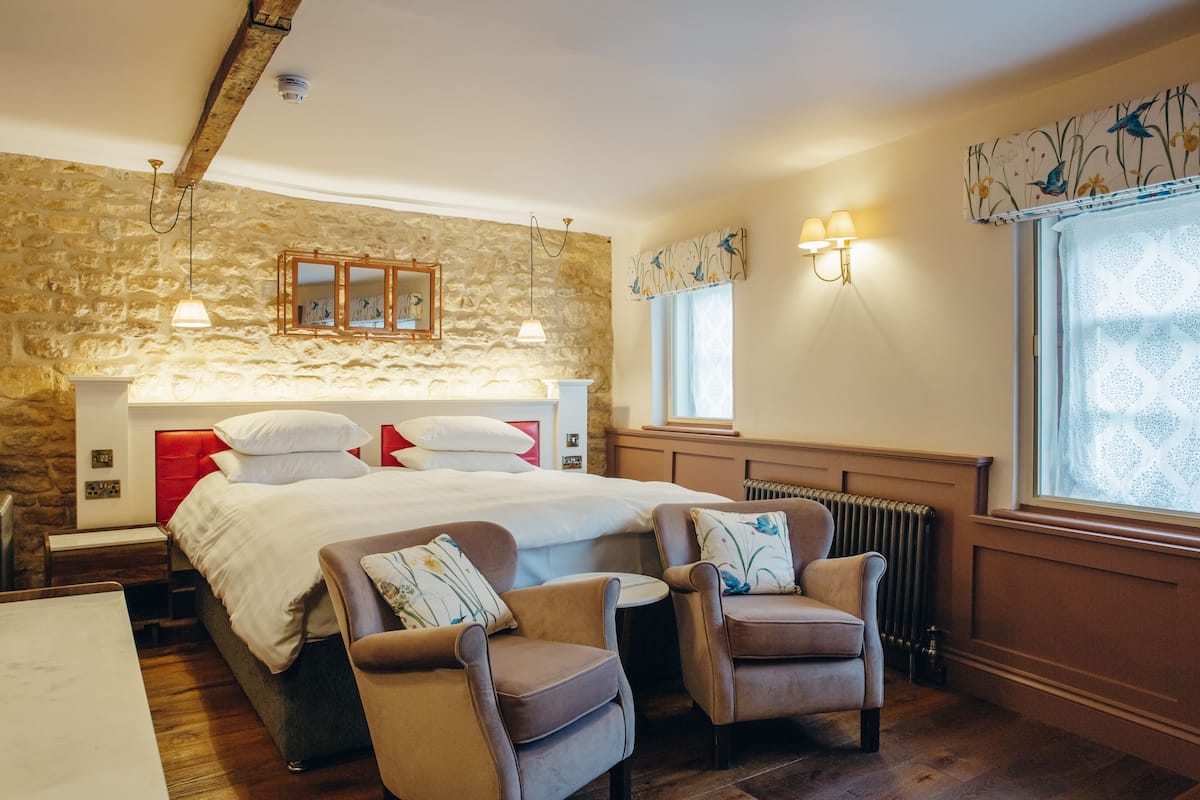 Een slaapkamer met een stenen muur, een bed met witte lakens, twee fauteuils, een houten hoofdbord en een raam met een bloemen gordijn.