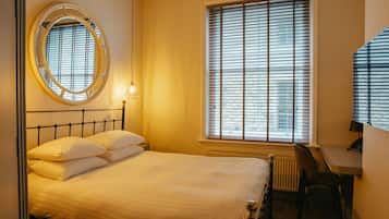 A neatly made bed with white linens, a round mirror on the wall, a desk with a chair, and a window with blinds.