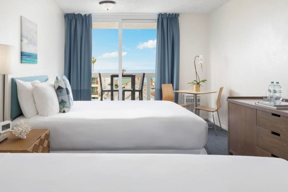 A hotel room with two beds, a desk, a chair, a table, a lamp, a plant, and a view of the sky and buildings.