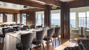Een eetgedeelte met meerdere tafels en stoelen, grote ramen met uitzicht op de oceaan, en een vluchtrouteborden boven de deur.
