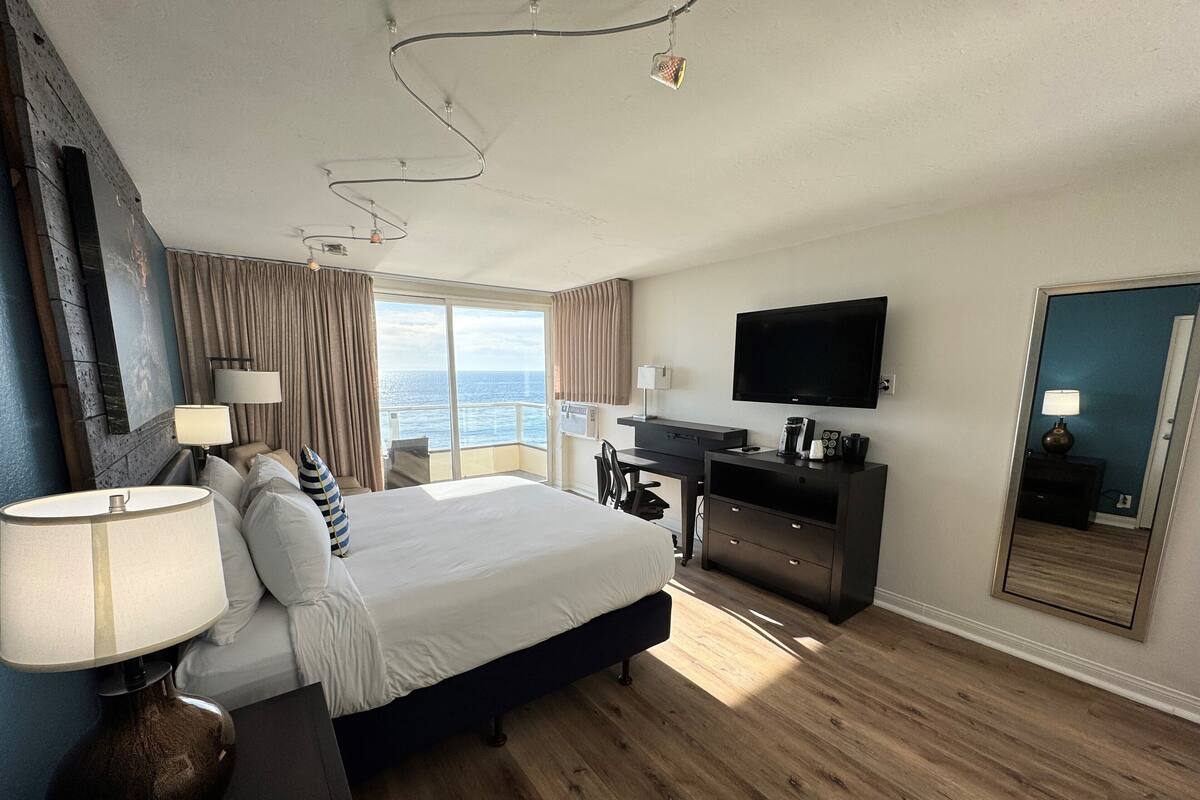 Une chambre d’hôtel avec un grand lit, un bureau équipé d’un téléviseur à écran plat, d’un ordinateur portable et d’une machine à café. Une porte coulissante donne sur le balcon, qui offre une vue sur la mer.