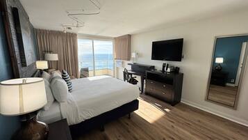 Habitación de hotel con una cama grande, un escritorio con televisión de pantalla plana, una computadora portátil y una cafetera. Tiene una puerta corredera al balcón con vistas al mar.
