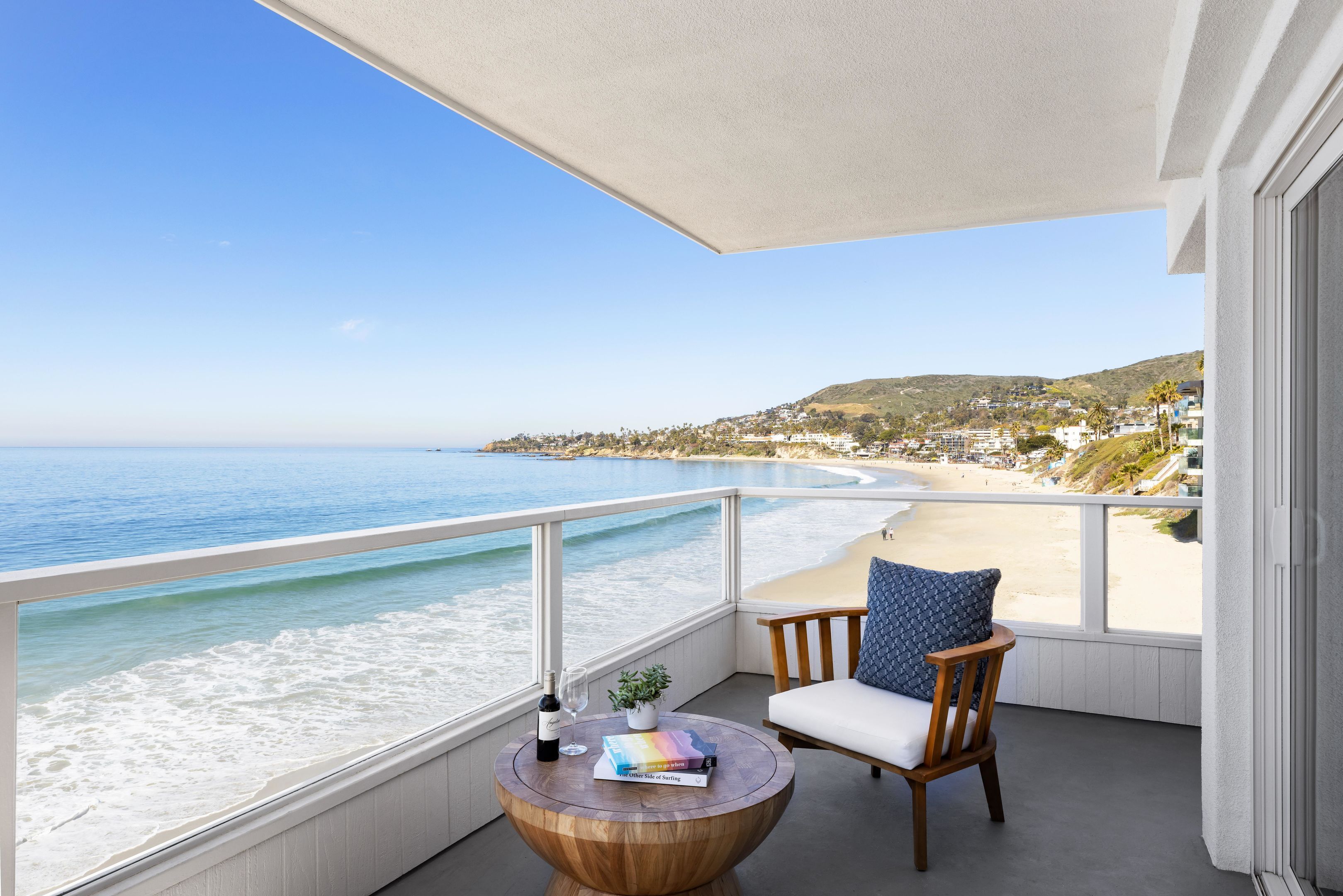 A balcony with a view of the beach, a wooden table, a chair, and a book.