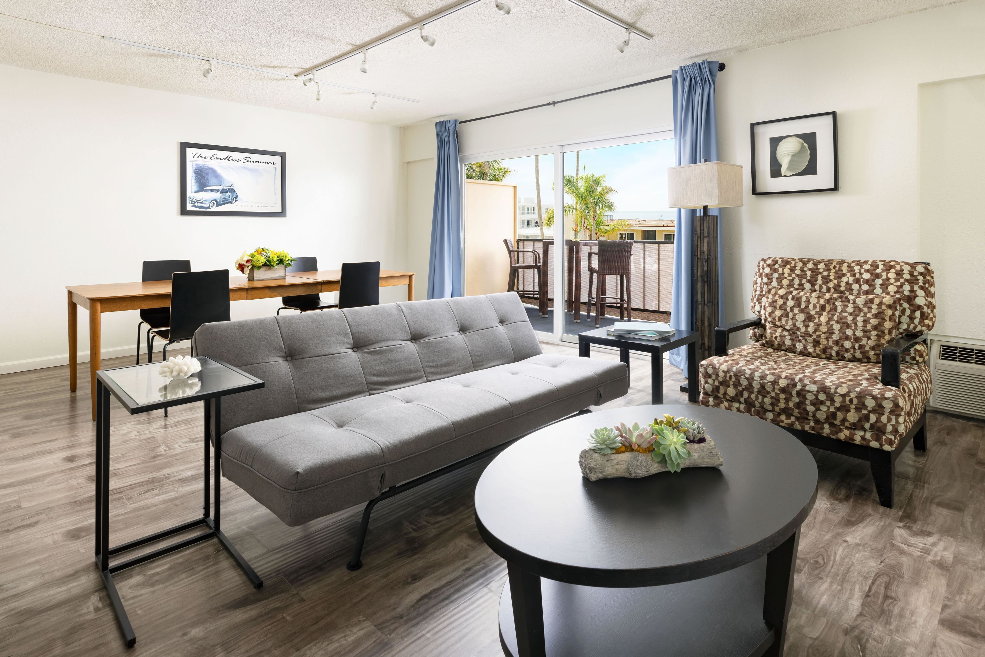 Una habitación de hotel moderna con un sofá gris, una mesa redonda de centro, un sillón estampado, una mesa de comedor con sillas y un ventanal con vistas a palmeras.