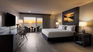 Een hotelkamer met een groot bed, een bureau met een flatscreen-tv, een stoel, een tafeltje en uitzicht op de ondergaande zon door de balkondeuren.