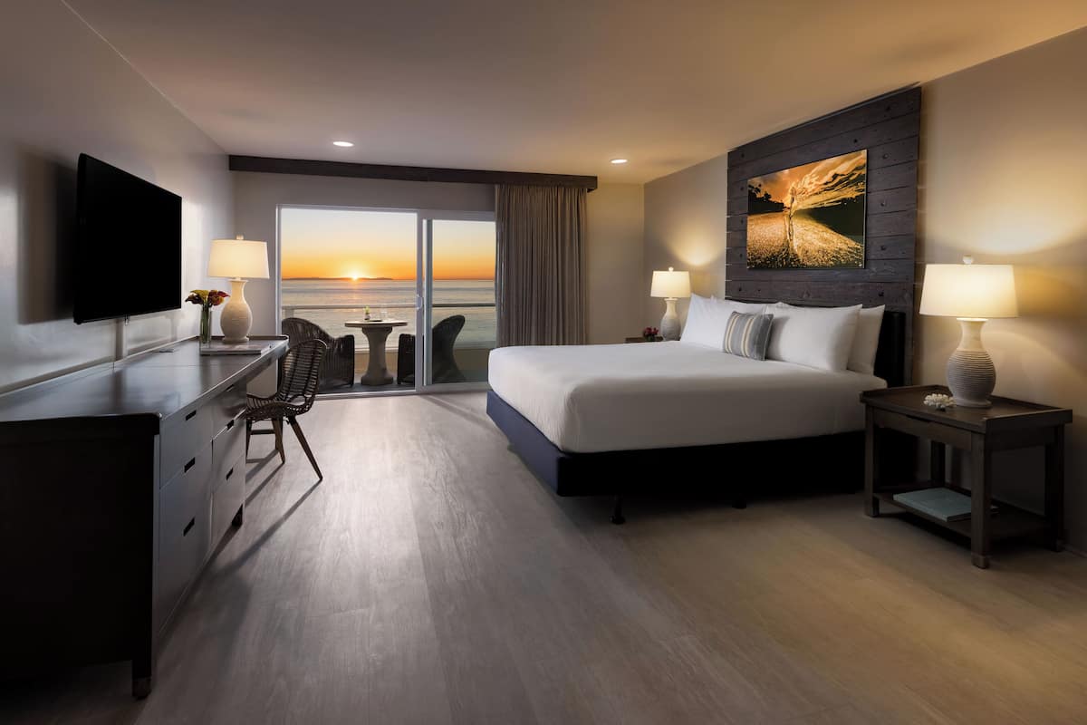 Une chambre d’hôtel avec un grand lit, un bureau doté d’un téléviseur à écran plat, une chaise, une petite table et une vue sur le coucher du soleil, visible à travers les portes du balcon.
