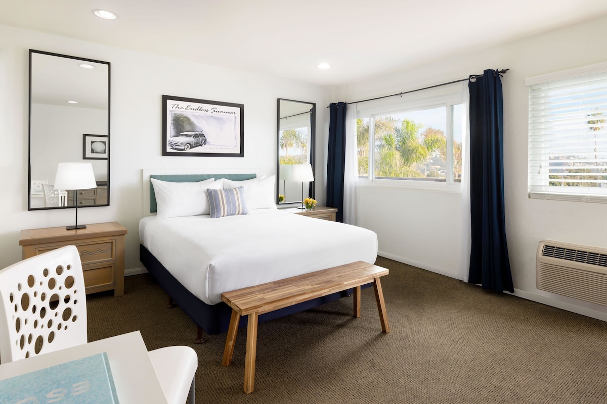 A hotel room with a bed, bedside tables, a bench, a large mirror, a window with curtains, and a view of palm trees.