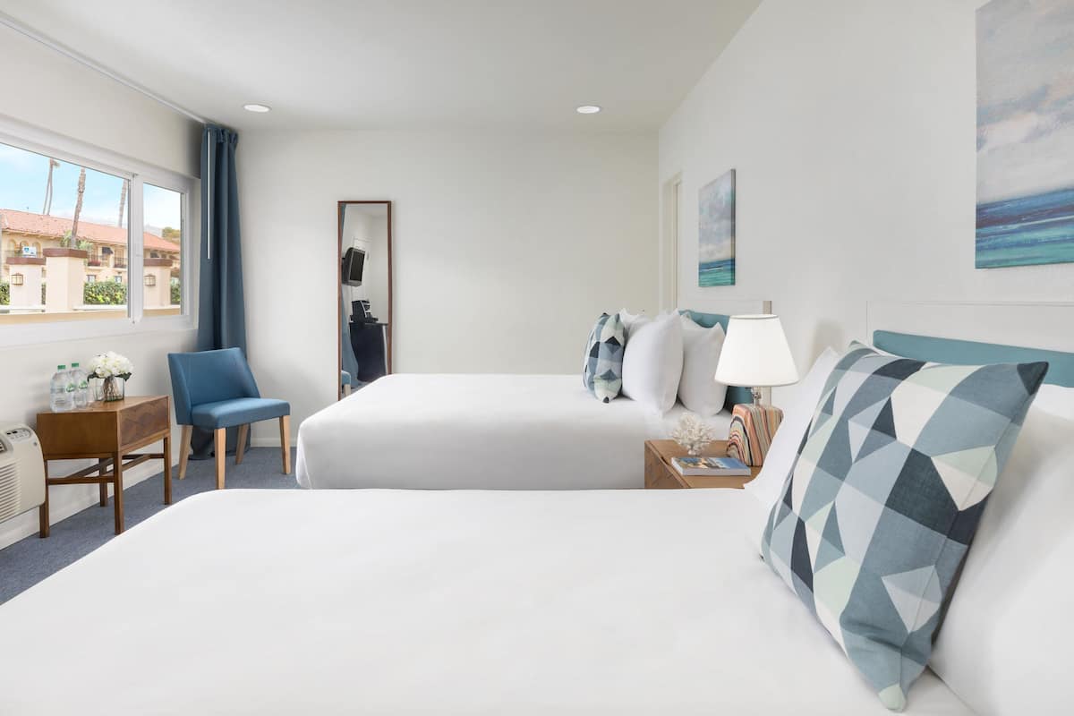 Habitación de hotel con dos camas, una silla, un escritorio con lámpara y ventana con vistas a edificios.