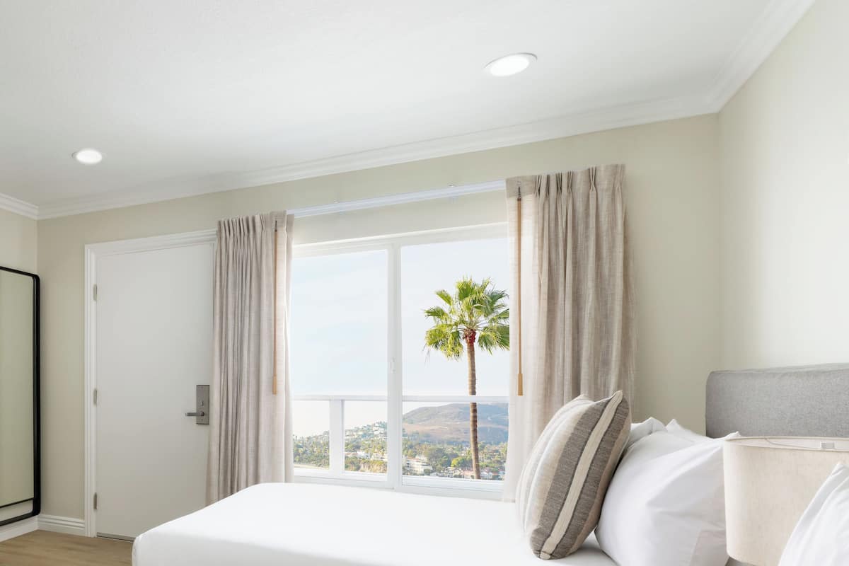Une chambre avec un grand lit, une vue sur un palmier et le paysage environnant, et une fenêtre avec des rideaux.